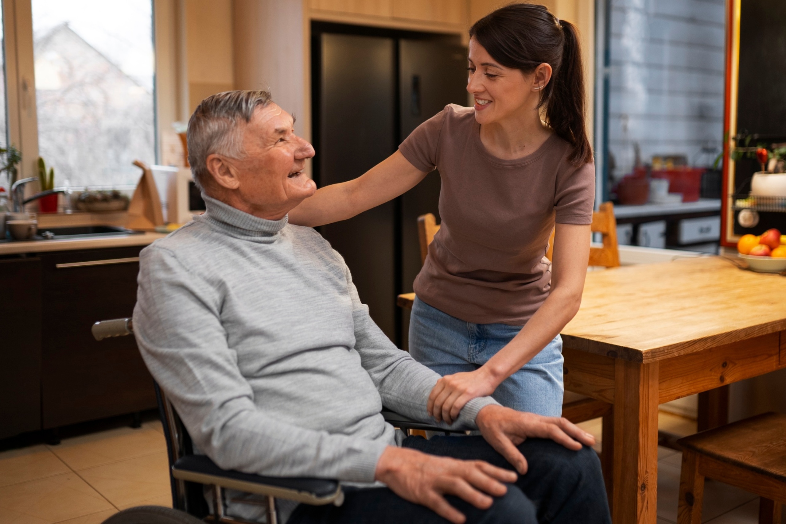 Young caregiver smiling and supporting an elderly man in a wheelchair at home, showing warmth and companionship.